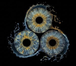 Vibrant grey-blue macro art of three human eye irises (brown, grey-blue, blue) merged with a dynamic water splash and droplet effect on a black background. This meaningful piece is part of the iris art online collection, symbolizing natural life and spiritual purity. Order your online iris scan now.